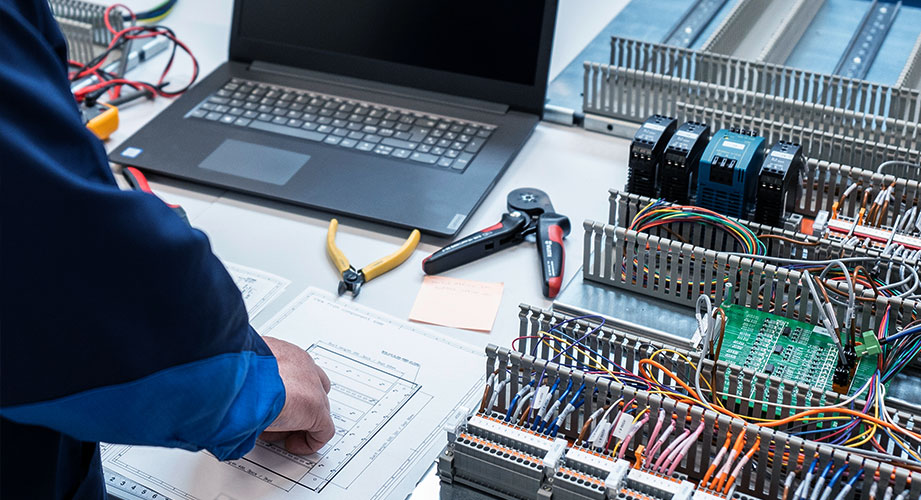 Työtekijä valmistaa koteloa, taustalla tietokone, piirustukset ja työvälineitä.