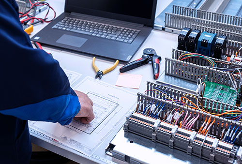 Työtekijä valmistaa koteloa, taustalla tietokone, piirustukset ja työvälineitä.